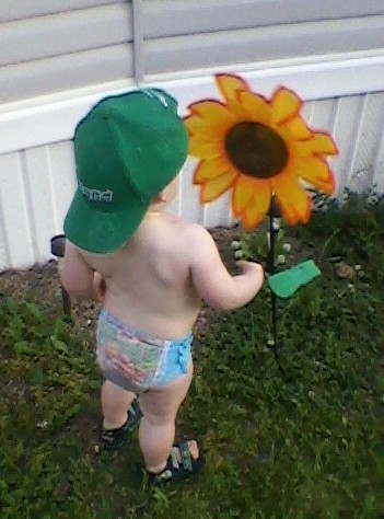 Photo of toddler boy, back turned to camera with green Irish hat turned around, looking at flower in front of a mobile home on a summer's day by Marion Weiscarger Rougsedge. Copyright 2012 - 2022, all rights reserved. Featured on Nippies.com Summer 2012 issue.